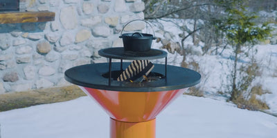 Video of a fire inside the orange Vuuris Tiger barbecue with a grill and pot on top on a snowy background with a stone house