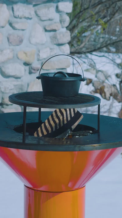 Video of a fire inside the orange Vuuris Tiger barbecue with a grill and pot on top on a snowy background with a stone house